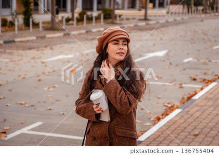 Woman in autumn colors enjoys hot coffee on a quiet city path. Soft wind, warm drink, and weekend simplicity. Woman in autumn colors enjoys hot coffee on a quiet city path. Soft wind, warm drink, and weekend simplicity. 136755404