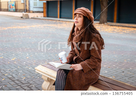 Side view of young pretty Caucasian woman in a coat and hat holds cup of coffee sitting on bench and holds book in city street. Lifestyle and city life Side view of young pretty Caucasian woman in a coat and hat holds cup of coffee sitting on bench and holds book in city street. Lifestyle and city life 136755405