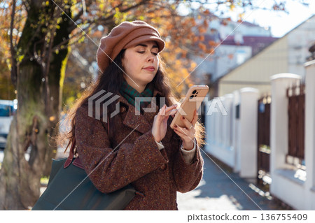 Mid shot of a young Caucasian woman in a autumn coat and beret using smartphone on street. City lifestyle 136755409