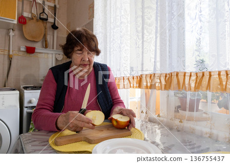 Elderly Caucasian woman cuts ripe apple on a kitchen table during daily meal preparation. She focuses on healthy eating and active aging. Concept of senior diet, prevention, and long life. 136755437
