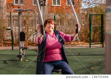 Elderly woman doing strength exercise on outdoor fitness equipment. Active lifestyle and senior wellness. Elderly woman doing strength exercise on outdoor fitness equipment. Active lifestyle and senior wellness. 136755449