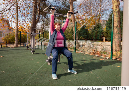 Older Caucasian woman using outdoor fitness machines in a green park. Physical activity, health, and long life in retirement. 136755459