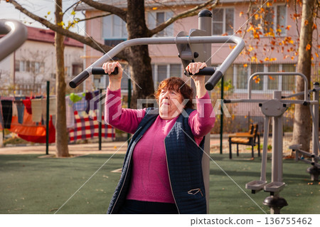 Senior Caucasian woman exercising on outdoor fitness equipment in a public park. Active retirement, healthy aging, and longevity lifestyle. 136755462