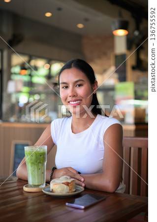 Smiling Woman Drinking Iced Matcha Latte at Cafe with Dessert 136755592