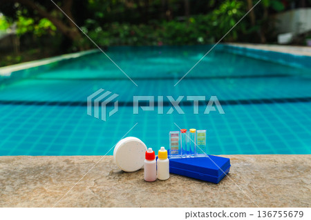 Pool water testing kit with chlorine tablet and reagent bottles on stone edge, tropical swimming pool background  136755679