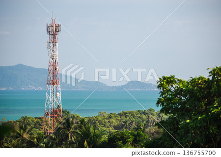 Mobile communication tower rising above tropical forest and turquoise sea with distant island mountains and wide blue sky copy space 136755700