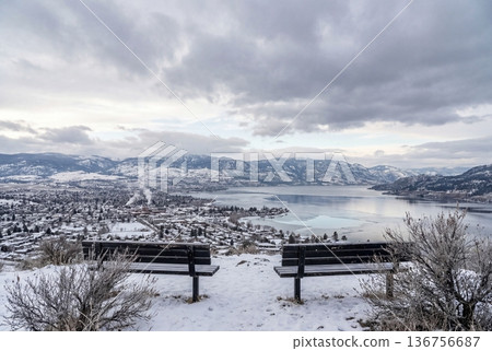 Two empty benches overlooking a snowy town and frozen lake surrounded by mountains under a cloudy sky 136756687