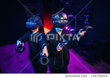 Cooperation, standing together. Two young men in virtual reality glasses indoors Cooperation, standing together. Two young men in virtual reality glasses indoors 136756939