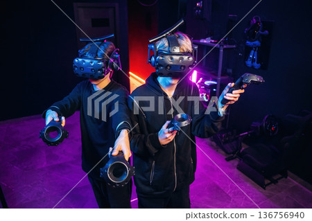 Cooperation, standing together. Two young men in virtual reality glasses indoors Cooperation, standing together. Two young men in virtual reality glasses indoors 136756940