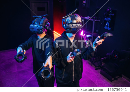 Cooperation, standing together. Two young men in virtual reality glasses indoors Cooperation, standing together. Two young men in virtual reality glasses indoors 136756941