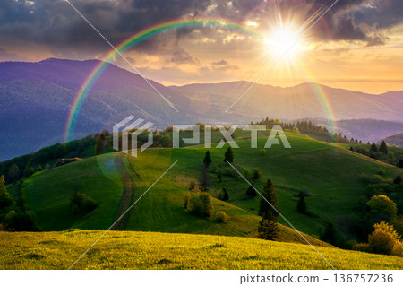 mountainous countryside scenery in springtime at sunset. dirt road and trees on the rolling hills in evening light. mountain ridge in the distance. alpine rural landscape. weather with rainbow 136757236