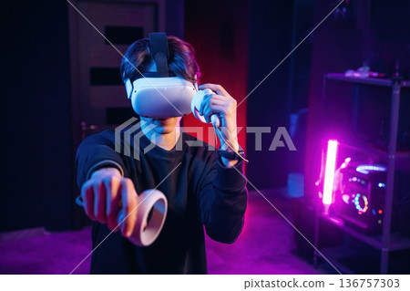 Interacting with game by using controllers. Young man is in virtual reality glasses indoors Interacting with game by using controllers. Young man is in virtual reality glasses indoors 136757303