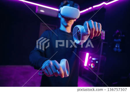 Interacting with game by using controllers. Young man is in virtual reality glasses indoors Interacting with game by using controllers. Young man is in virtual reality glasses indoors 136757307