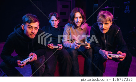 Looking forward, playing together. Young gamers are with console controllers indoors Looking forward, playing together. Young gamers are with console controllers indoors 136757355