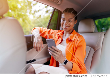 Young black woman sitting in car back seat smiling and browsing smartphone Young black woman sitting in car back seat smiling and browsing smartphone 136757453