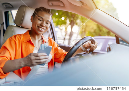 Smiling young black woman driving car and looking at smartphone in hand 136757464