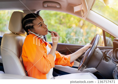 Young black woman applying lip gloss and talking on cellphone while driving 136757471