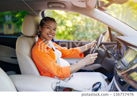 Smiling pregnant black woman driving car on sunny day, wearing seatbelt 136757476