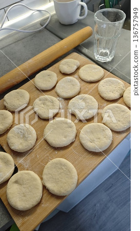 Raw dough pieces on wooden board with rolling pin and glass of water in kitchen. Home baking process and preparation concept Raw dough pieces on wooden board with rolling pin and glass of water in kitchen. Home baking process and preparation concept 136757949