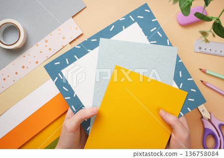 Hands holding colored paper for crafts. Tape, scissors, pencil on beige background. DIY handmade project for children. Hands holding colored paper for crafts. Tape, scissors, pencil on beige background. DIY handmade project for children. 136758084
