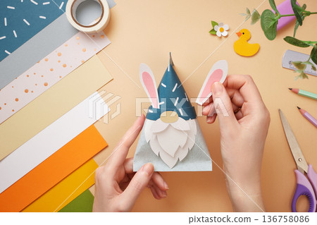 Hands attach rabbit ears to paper gnome. Tape, scissors, pencil on beige background. DIY handmade project for children. Hands attach rabbit ears to paper gnome. Tape, scissors, pencil on beige background. DIY handmade project for children. 136758086