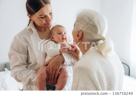 Standing, looking at each other. Mother, grandmother and little baby child on hands indoors Standing, looking at each other. Mother, grandmother and little baby child on hands indoors 136758249