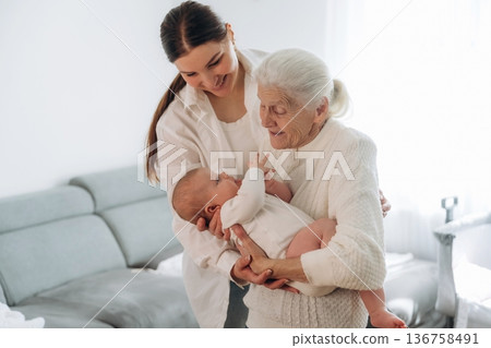 Cheerful facial expression. Mother, grandmother and little baby child on hands indoors Cheerful facial expression. Mother, grandmother and little baby child on hands indoors 136758491