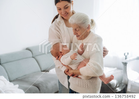 Cheerful facial expression. Mother, grandmother and little baby child on hands indoors Cheerful facial expression. Mother, grandmother and little baby child on hands indoors 136758492