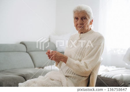 Looking at the camera. Grandma sits in a chair at home and knits clothes Looking at the camera. Grandma sits in a chair at home and knits clothes 136758647