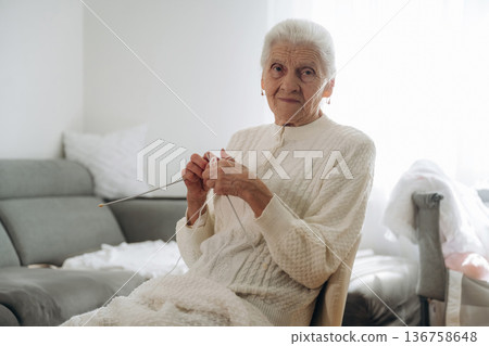 Looking at the camera. Grandma sits in a chair at home and knits clothes 136758648