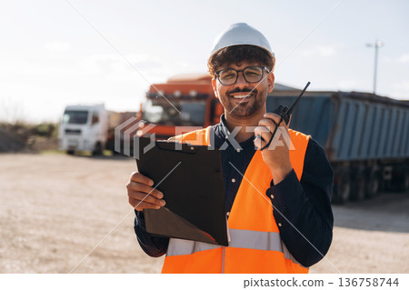 In white hard hat and with walkie talkie. Male worker is near truck outdoors In white hard hat and with walkie talkie. Male worker is near truck outdoors 136758744