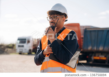 Walkie talkie in hand, talking. Male worker is near truck outdoors 136758894