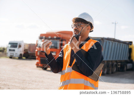 Walkie talkie in hand, talking. Male worker is near truck outdoors Walkie talkie in hand, talking. Male worker is near truck outdoors 136758895