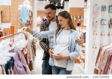 Feeling good, choosing the wear. Pregnant woman with man are in the kid's clothing store Feeling good, choosing the wear. Pregnant woman with man are in the kid's clothing store 136759469