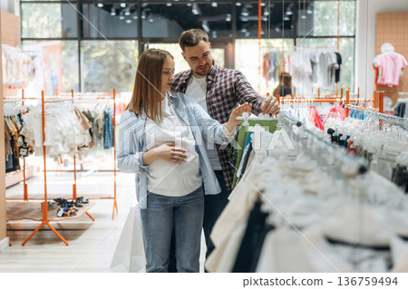 Side view, wear is hanging. Pregnant woman with man are in the kid's clothing store Side view, wear is hanging. Pregnant woman with man are in the kid's clothing store 136759494