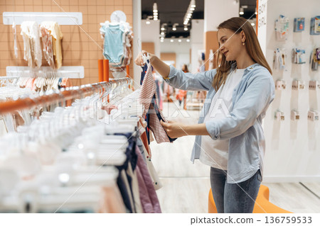 The cute little wear. Young pregnant woman is in the kid's clothing store The cute little wear. Young pregnant woman is in the kid's clothing store 136759533