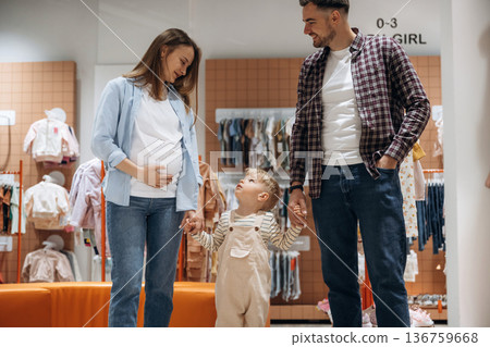 Walking and holding child by hands. Father, son and pregnant mother are in kid's clothing store 136759668