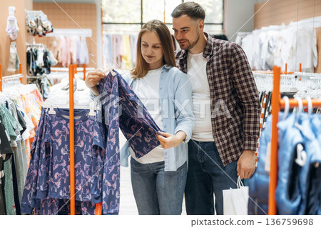 By the sweaters. Pregnant woman with man are in the kid's clothing store By the sweaters. Pregnant woman with man are in the kid's clothing store 136759698