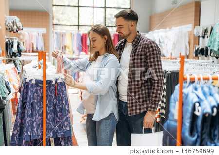 By the sweaters. Pregnant woman with man are in the kid's clothing store By the sweaters. Pregnant woman with man are in the kid's clothing store 136759699