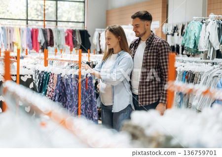 By the sweaters. Pregnant woman with man are in the kid's clothing store By the sweaters. Pregnant woman with man are in the kid's clothing store 136759701