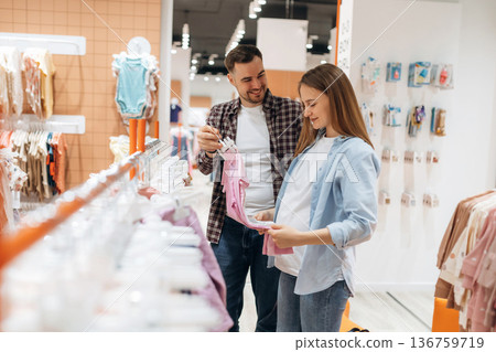 Clothes hanging in a row. Pregnant woman with man are in the kid's store 136759719