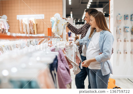 Clothes hanging in a row. Pregnant woman with man are in the kid's store Clothes hanging in a row. Pregnant woman with man are in the kid's store 136759722