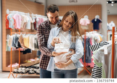 White cute little shoes. Pregnant woman with man are in the kid's clothing store 136759744
