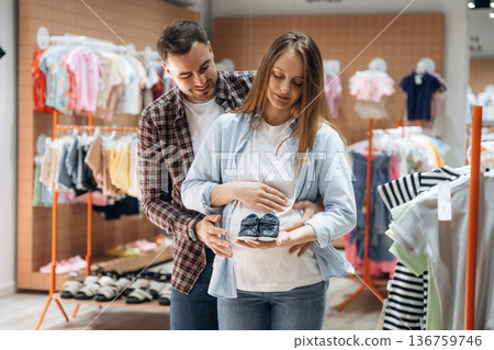 Embracing, holding little shoes. Pregnant woman with man are in the kid's clothing store Embracing, holding little shoes. Pregnant woman with man are in the kid's clothing store 136759746