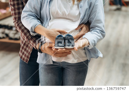 Embracing, holding little shoes. Pregnant woman with man are in the kid's clothing store 136759747