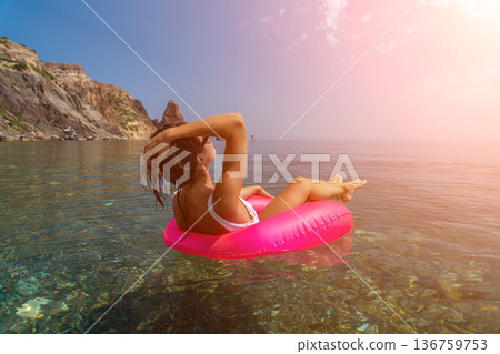 Woman floating clear water, relaxing on pink inflatable ring near stunning mediterranean cliffs enjoying tranquil summer vacation Woman floating clear water, relaxing on pink inflatable ring near stunning mediterranean cliffs enjoying tranquil summer vacation 136759753