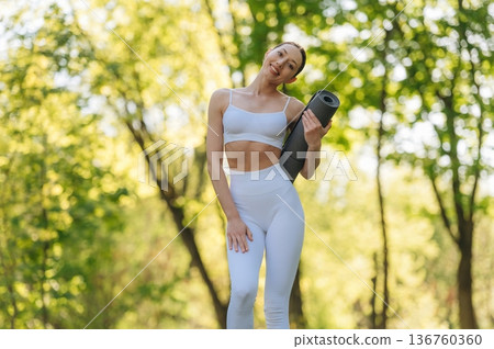 Front view. Standing with yoga mat. Beautiful fitness woman is outdoors, amazing nature Front view. Standing with yoga mat. Beautiful fitness woman is outdoors, amazing nature 136760360