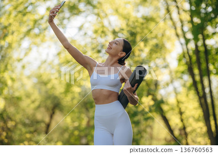 Smartphone and yoga mat are in hands. Beautiful fitness woman is outdoors, amazing nature 136760563
