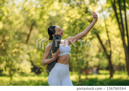 Smartphone and yoga mat are in hands. Beautiful fitness woman is outdoors, amazing nature 136760564