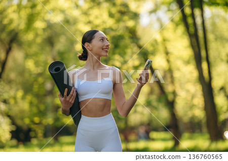 Smartphone and yoga mat are in hands. Beautiful fitness woman is outdoors, amazing nature Smartphone and yoga mat are in hands. Beautiful fitness woman is outdoors, amazing nature 136760565